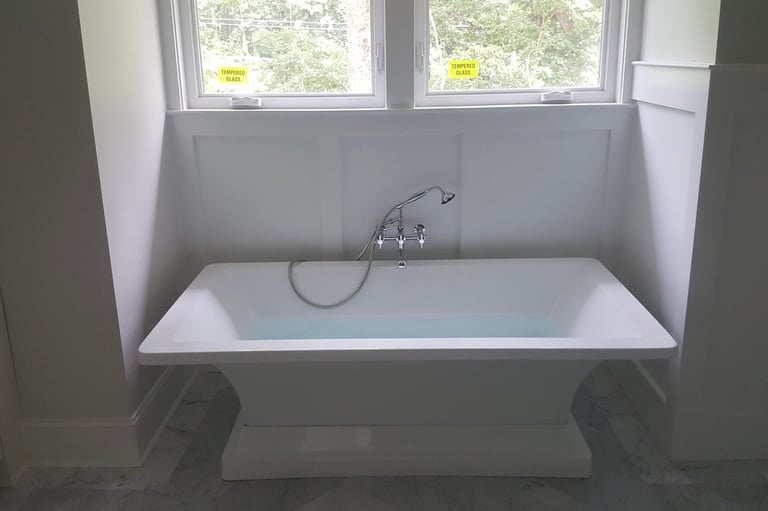Modern white rectangular bathtub with chrome faucet positioned beneath two windows in a minimalist bathroom