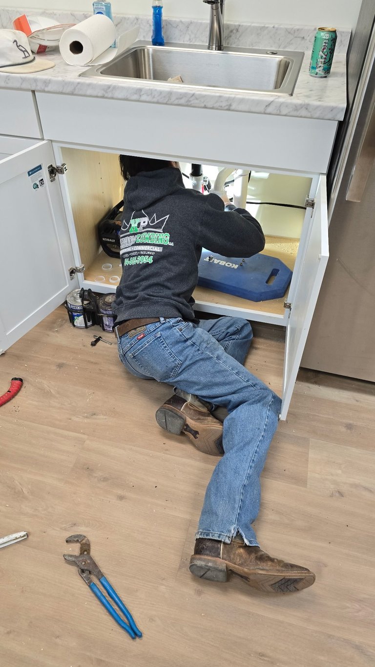 Plumber working under kitchen sink cabinet with tools, fixing pipes and plumbing fixtures.