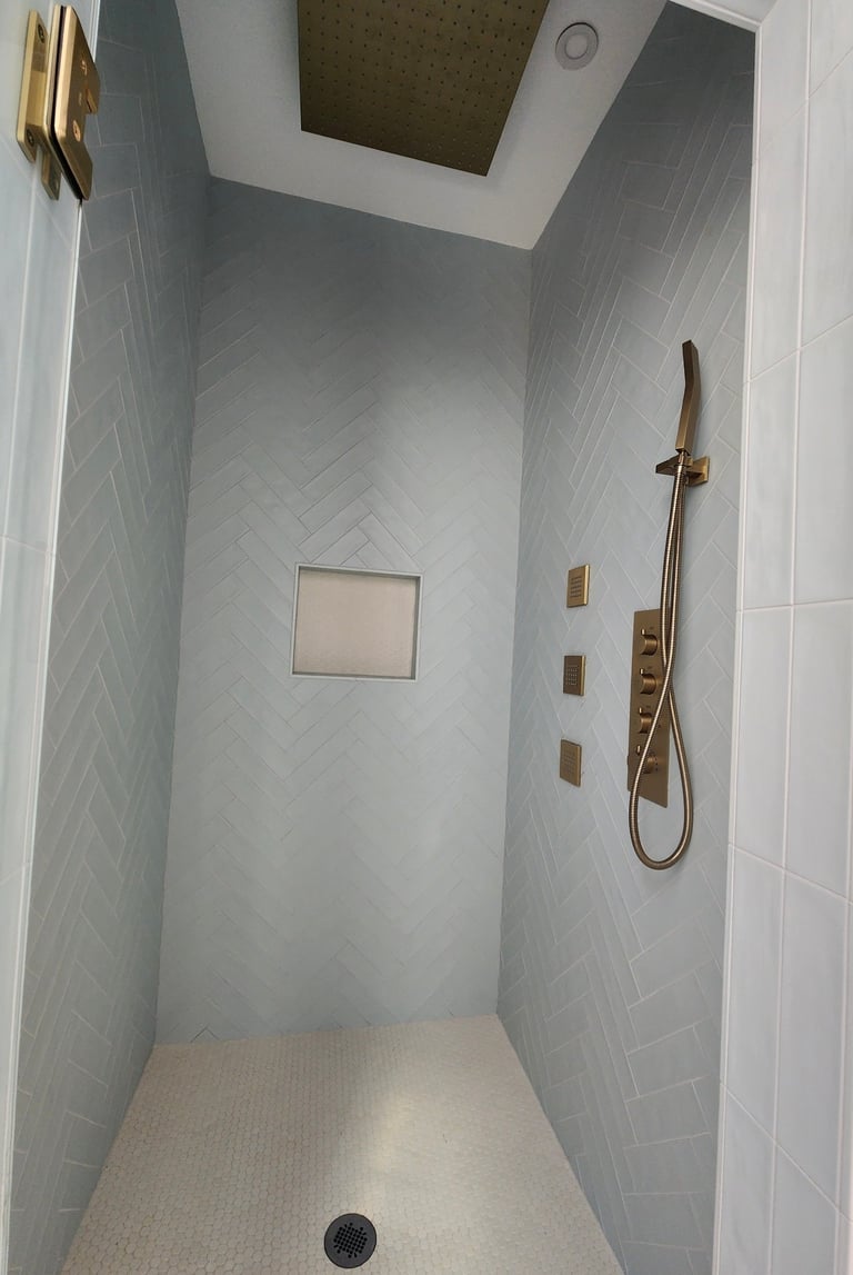 Modern walk-in shower with light gray herringbone tile walls, brass fixtures, rainfall showerhead, and built-in shelving niche.