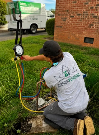 HVAC technician in white shirt kneeling outdoors reading gauges on air conditioning unit with service van in background