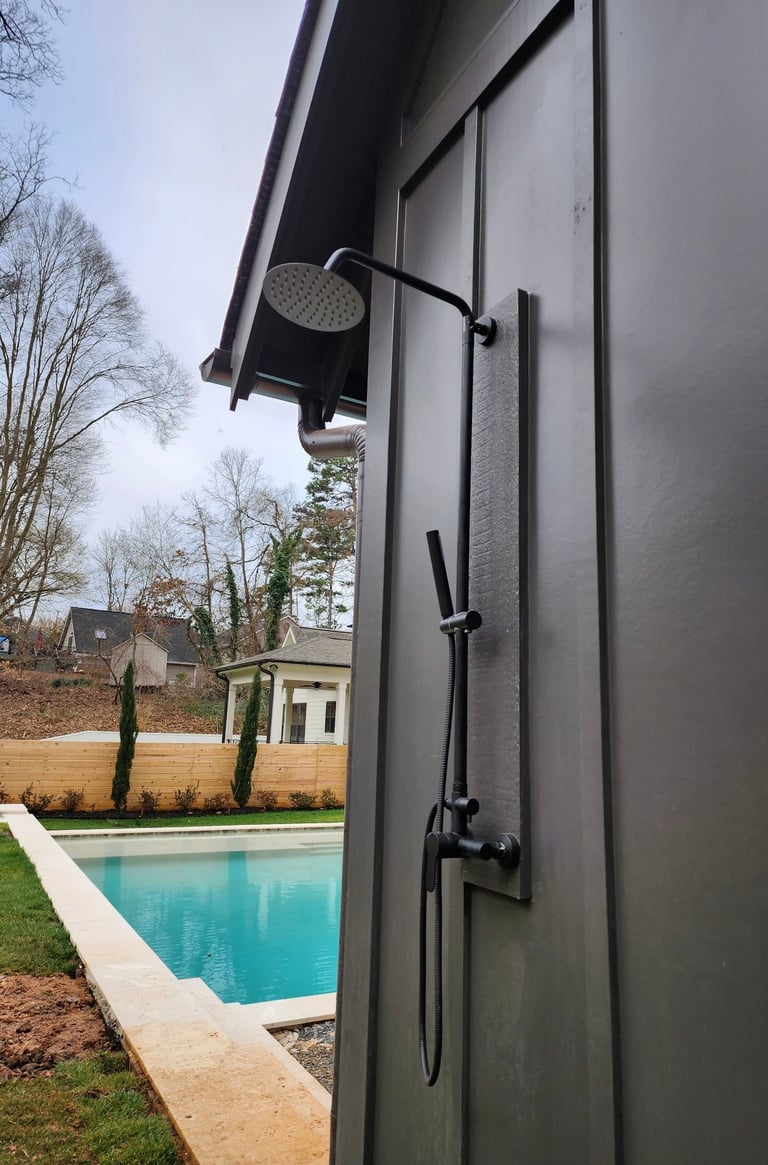 Modern black outdoor shower with rainfall and handheld showerheads mounted on gray wall overlooking a swimming pool